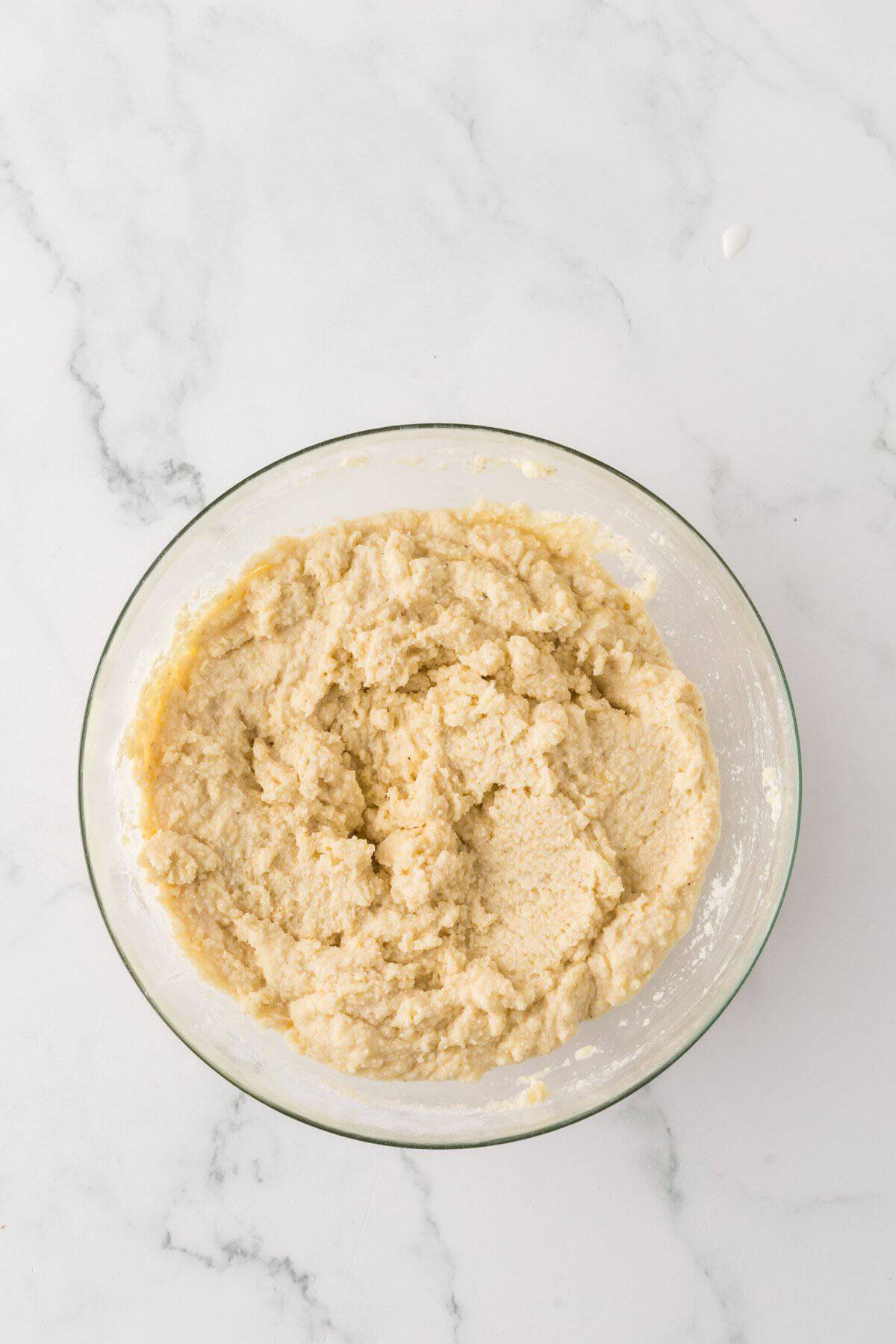 A clear glass bowl filled with thick, creamy cookie dough is placed on a white marble countertop. The dough has a light beige color and a slightly rough texture, indicating the mixture of flour, sugar, butter, and other ingredients.