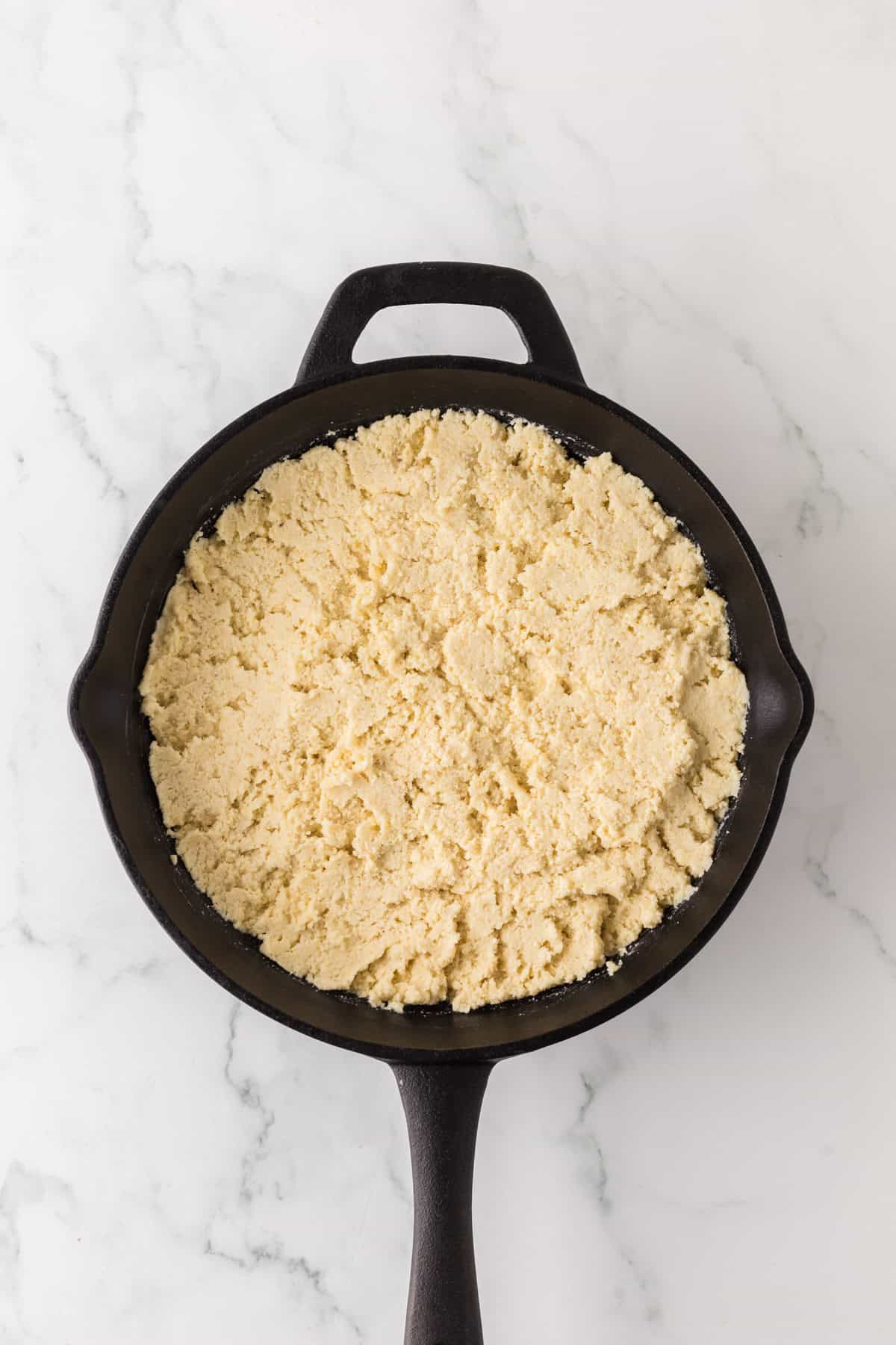 A cast iron skillet filled with crumbly, pale yellow topping sits on a white marble countertop.