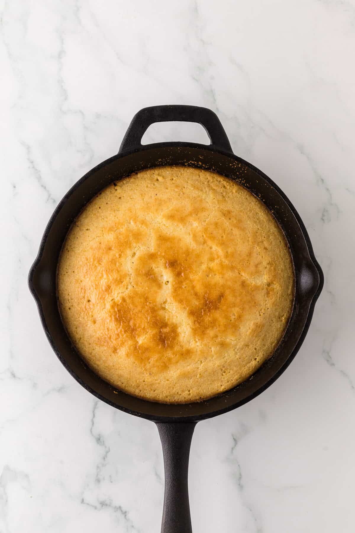 A golden-brown cornbread is baked in a round cast iron skillet. The skillet is placed on a white marble surface. The cornbread has a slightly domed surface and an even, crisp texture.