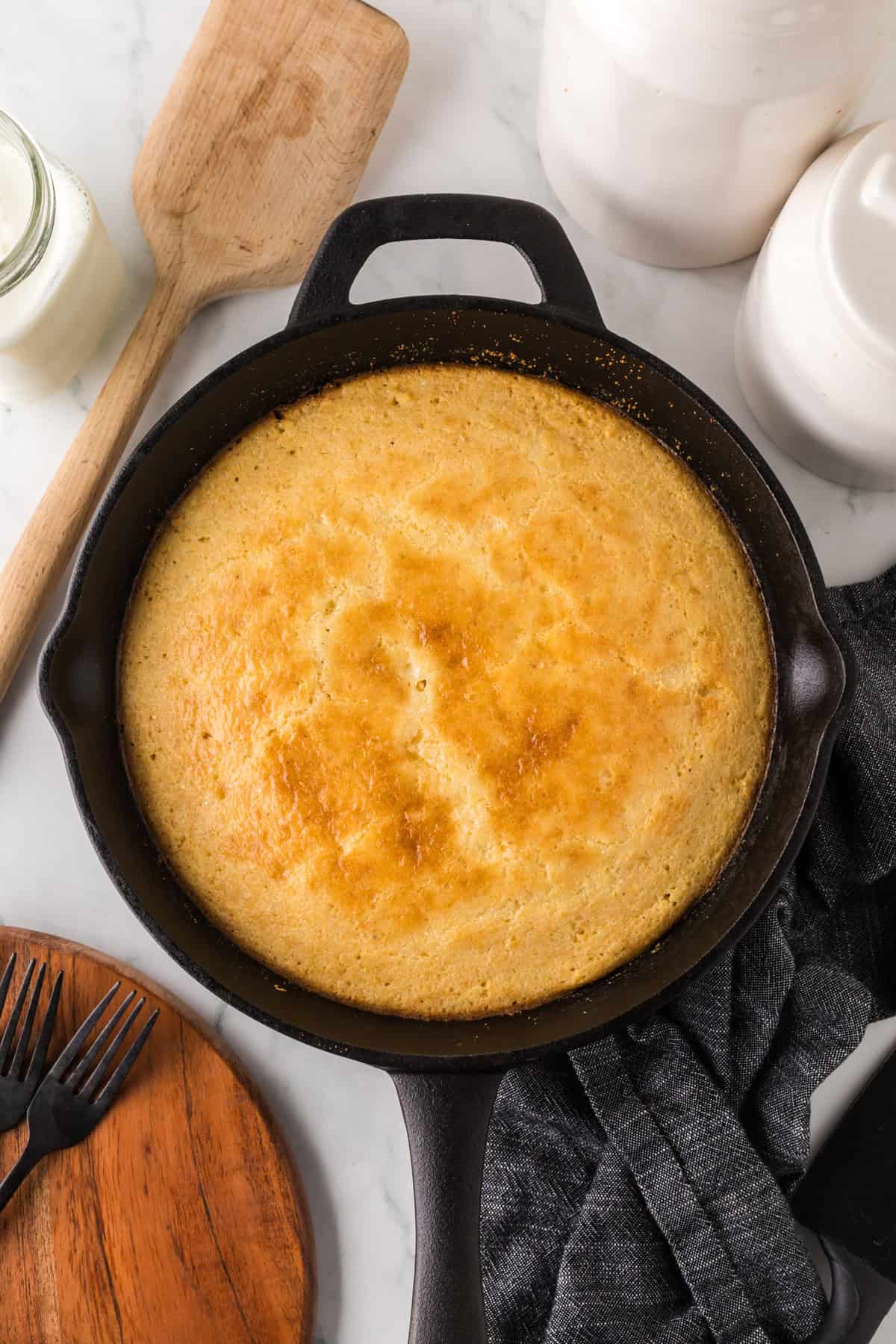 A golden-brown cornbread in a cast-iron skillet on a marble countertop. Nearby are a wooden spatula, a gray cloth, and black forks on a wooden board. White jars and a glass of milk are in the background.