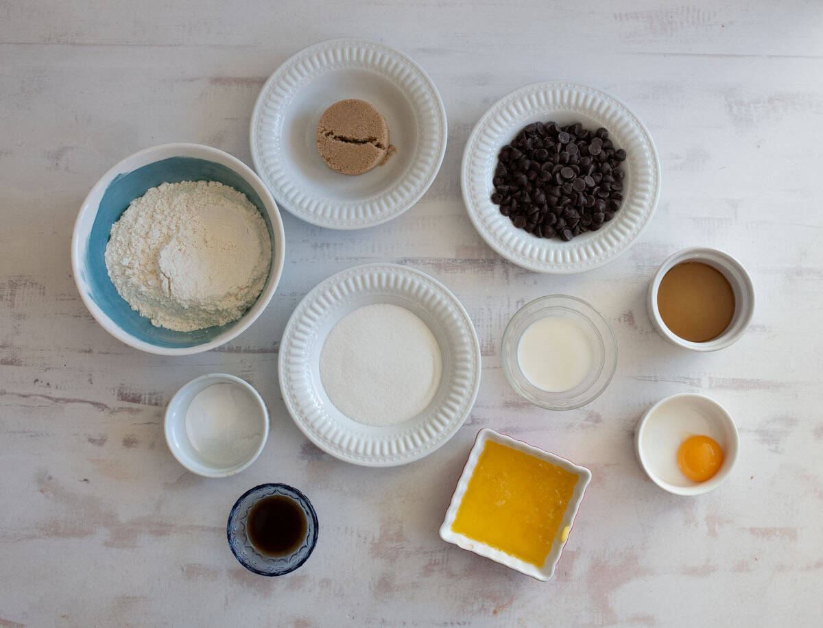 Various baking ingredients are arranged on a white surface, including flour, brown sugar, chocolate chips, granulated sugar, butter, an egg, vanilla extract, milk, and a small bowl with a liquid ingredient.