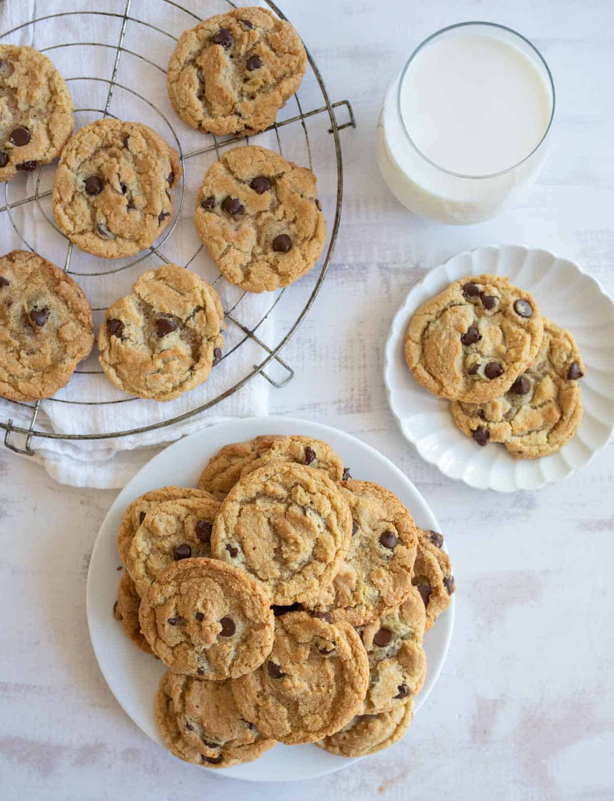 A plate piled with chocolate chip cookies sits on a white surface, next to a smaller plate with two cookies. A cooling rack holds more cookies above. A glass of milk is nearby, completing the cozy dessert setting.