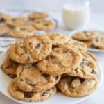 A plate of freshly baked thin and crispy chocolate chip cookies is stacked in the foreground, with more cookies on a rack and a glass of milk in the background. The scene suggests a cozy, homemade treat.