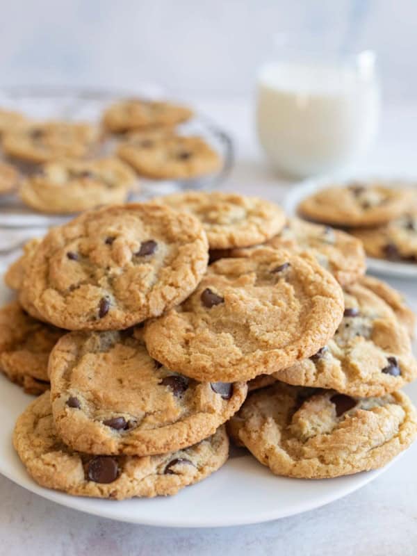 A plate of freshly baked thin and crispy chocolate chip cookies is stacked in the foreground, with more cookies on a rack and a glass of milk in the background. The scene suggests a cozy, homemade treat.