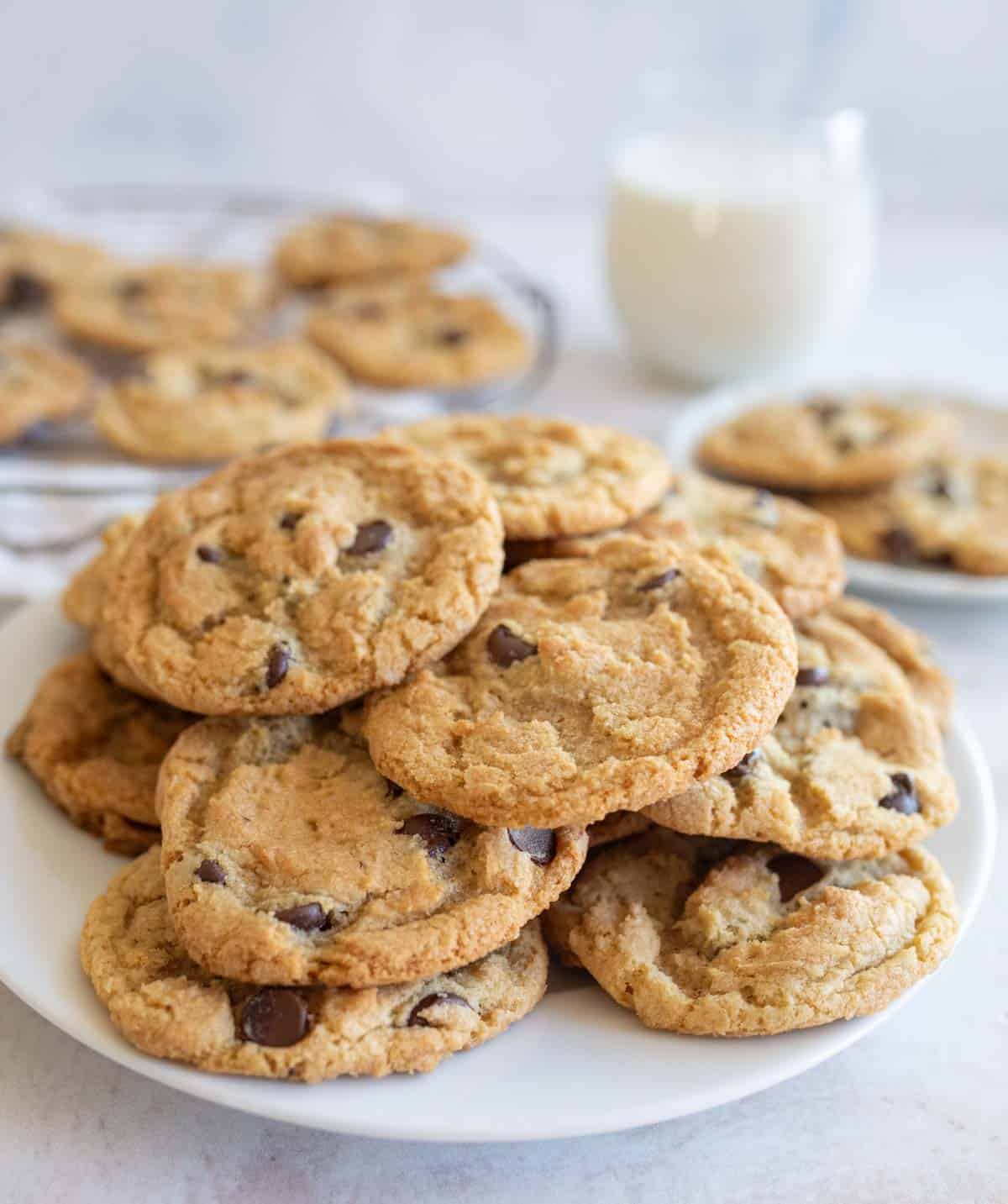 A plate of freshly baked thin and crispy chocolate chip cookies is stacked in the foreground, with more cookies on a rack and a glass of milk in the background. The scene suggests a cozy, homemade treat.