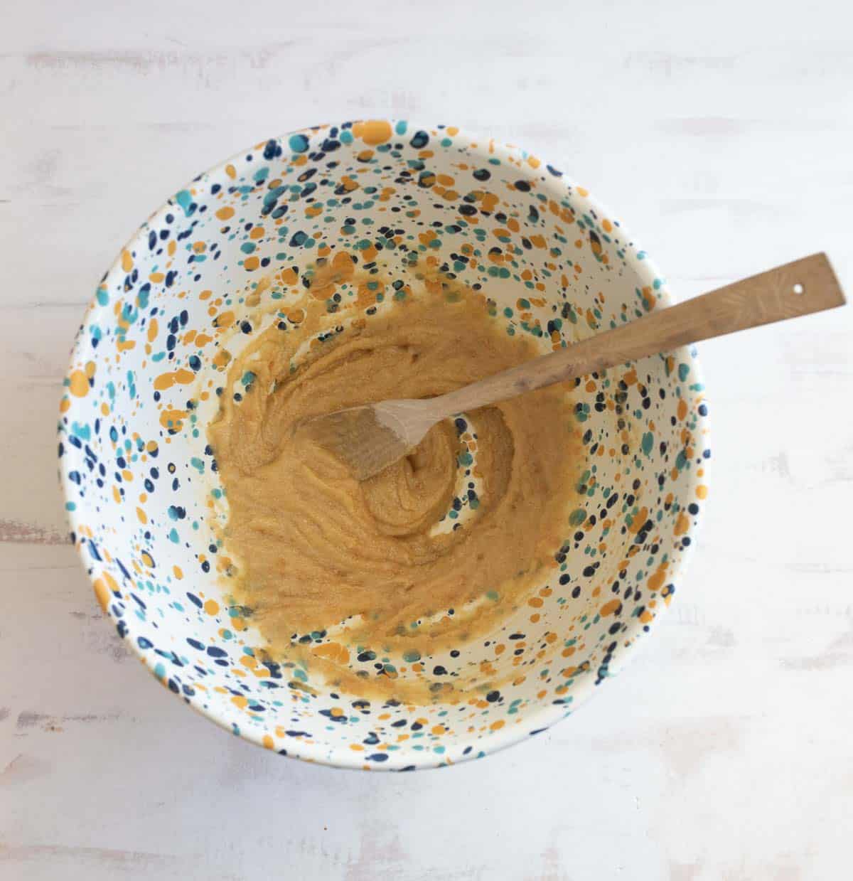 A speckled mixing bowl filled with a creamy batter sits on a light, textured surface. A wooden spoon rests in the bowl, partially covered by the light brown batter.