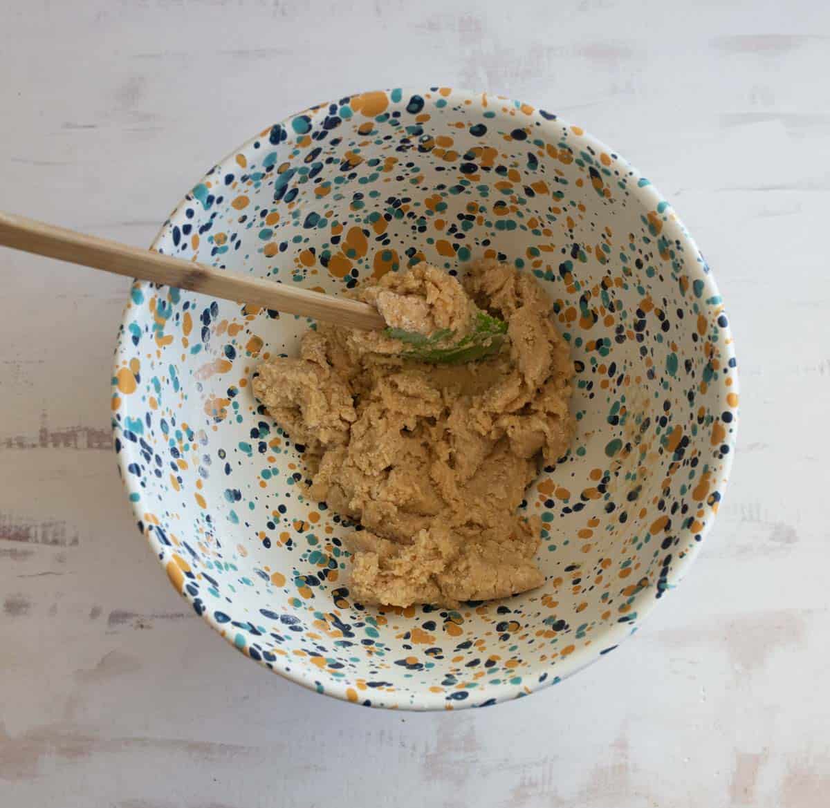 A colorful speckled bowl with a sticky dough and a wooden spatula. The bowls interior is white with blue, yellow, and orange speckles. The dough appears thick and slightly textured. The background is a distressed white surface.