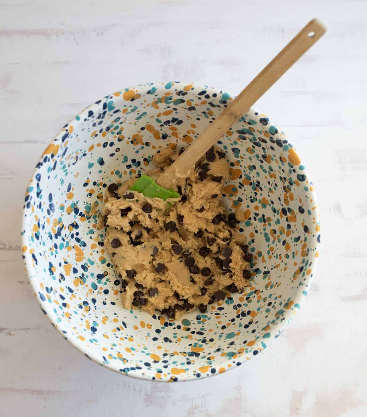 A ceramic bowl with a colorful splatter pattern holds cookie dough mixed with chocolate chips. A wooden spoon with a green handle is placed in the dough. The bowl is on a light-colored surface.