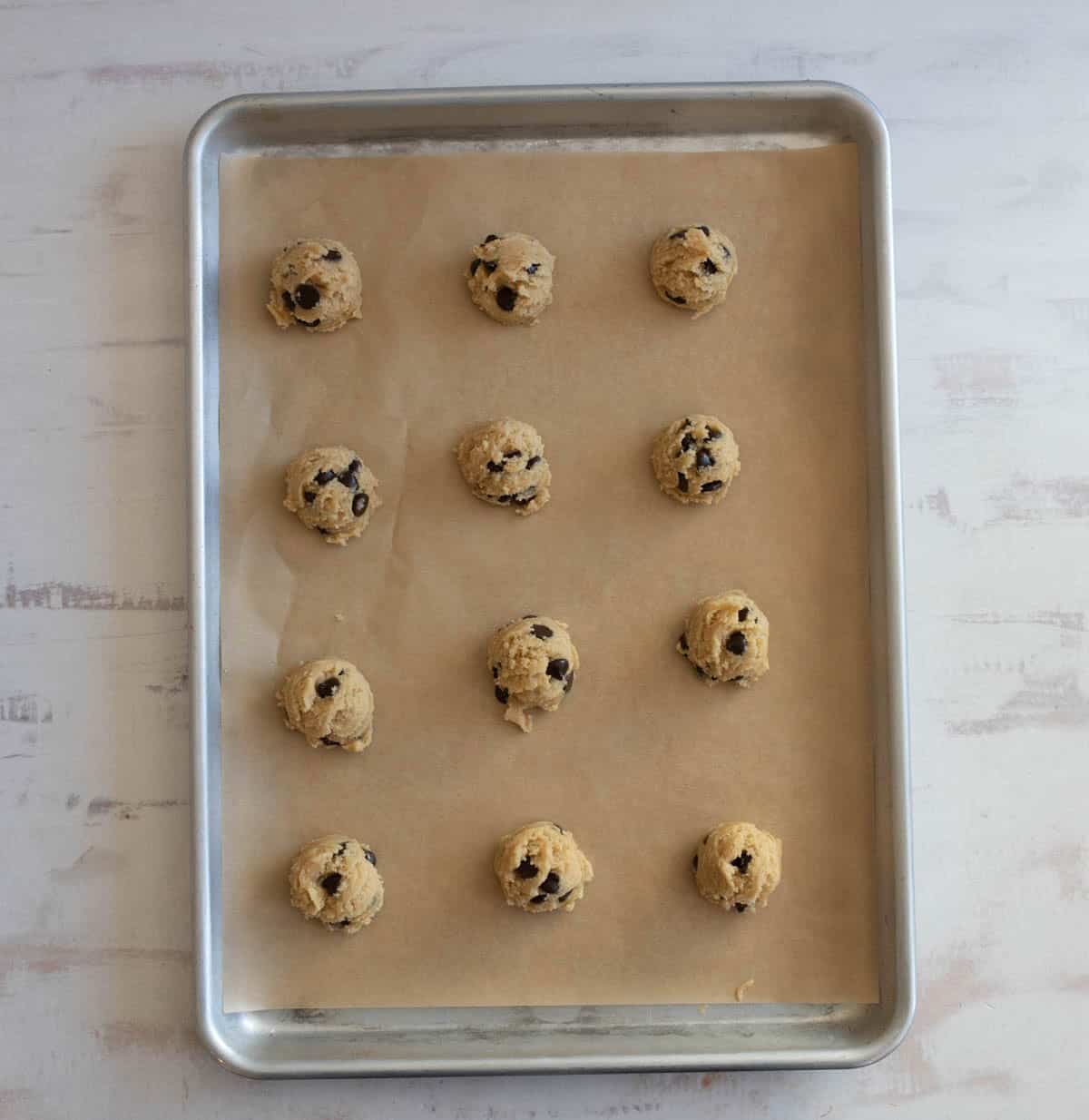 Twelve balls of chocolate chip cookie dough are evenly spaced on a parchment-lined baking sheet, ready to be baked. The baking sheet is placed on a light-colored wooden surface.