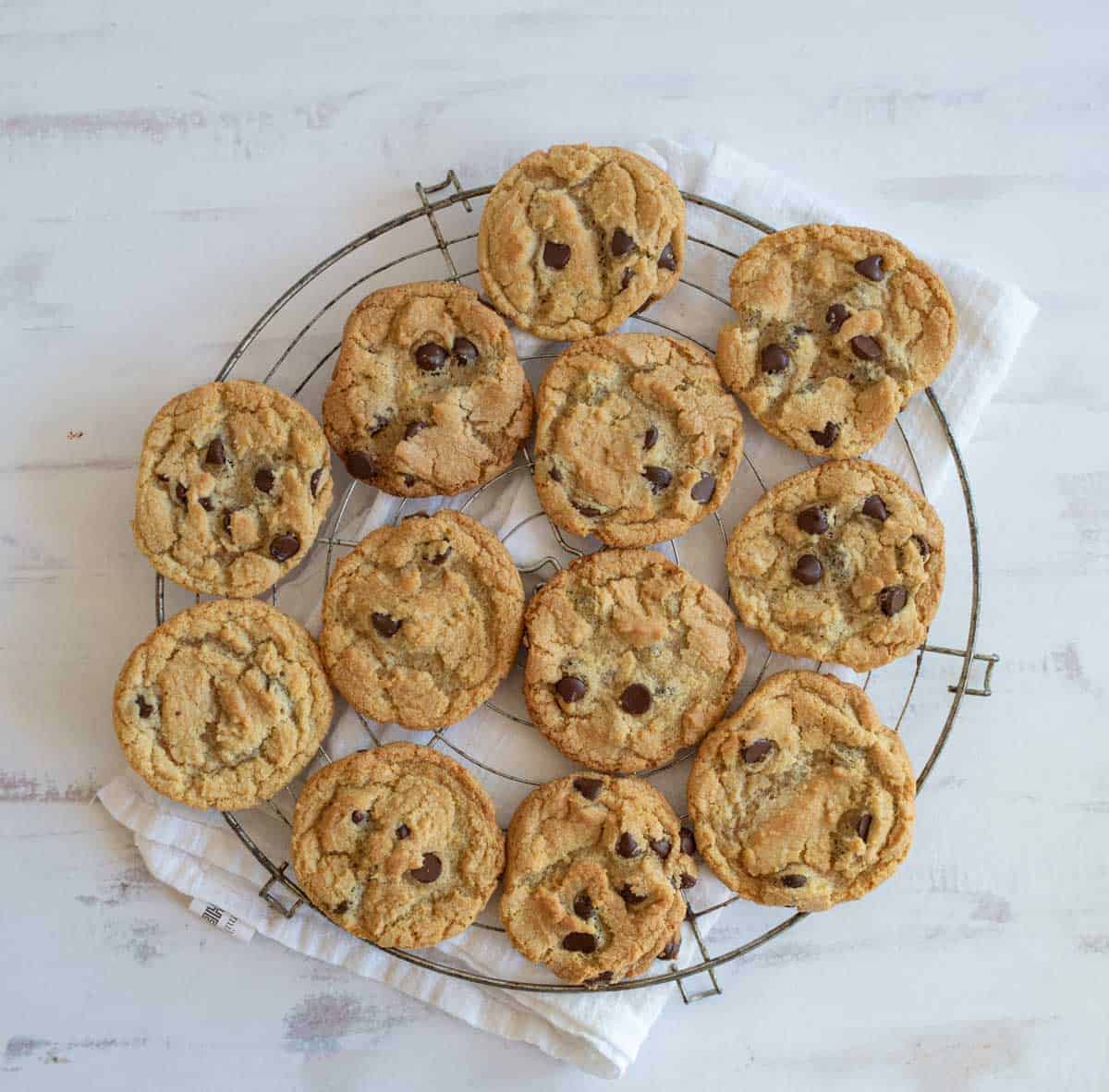 A cooling rack holds 12 freshly baked chocolate chip cookies on a light-colored cloth. The cookies are golden brown with visible chocolate chips, arranged in a circular pattern on a white wooden surface.