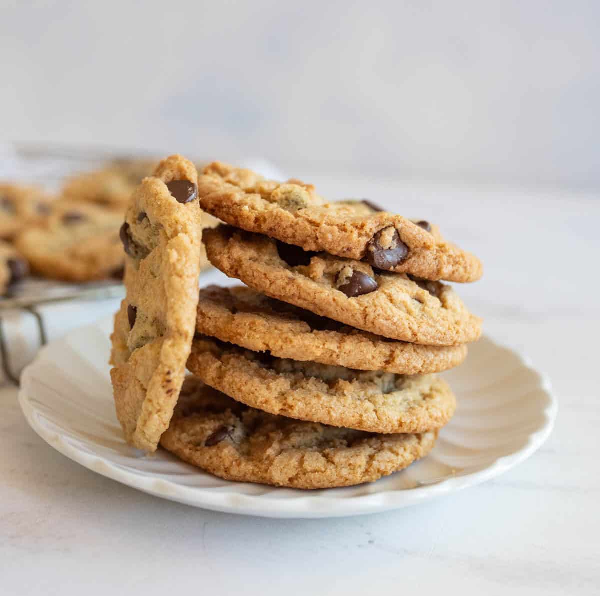 A stack of chocolate chip cookies on a white plate, set against a light-colored background. The cookies are golden brown with visible chocolate chips, suggesting a freshly baked texture.