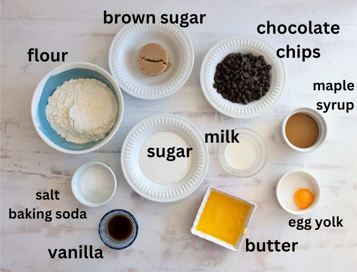 Ingredients for baking are arranged in bowls on a white surface: flour, brown sugar, chocolate chips, maple syrup, milk, sugar, an egg yolk, butter, vanilla, salt, and baking soda. Each is labeled with text.