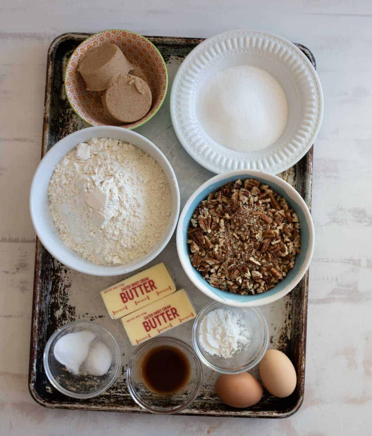 Baking ingredients arranged on a tray include two sticks of butter, bowls of flour, pecans, white and brown sugar, a small bowl of salt, a glass bowl with vanilla extract, and two eggs.