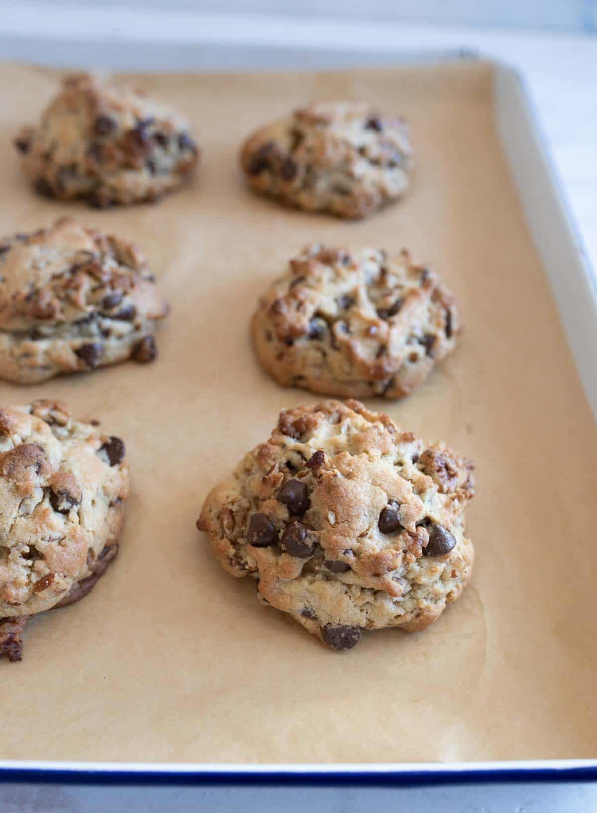 A baking tray with six freshly baked chocolate chip cookies on parchment paper. The cookies are golden-brown and chunky, with visible chocolate chips. The background is slightly blurred, focusing on the cookies.