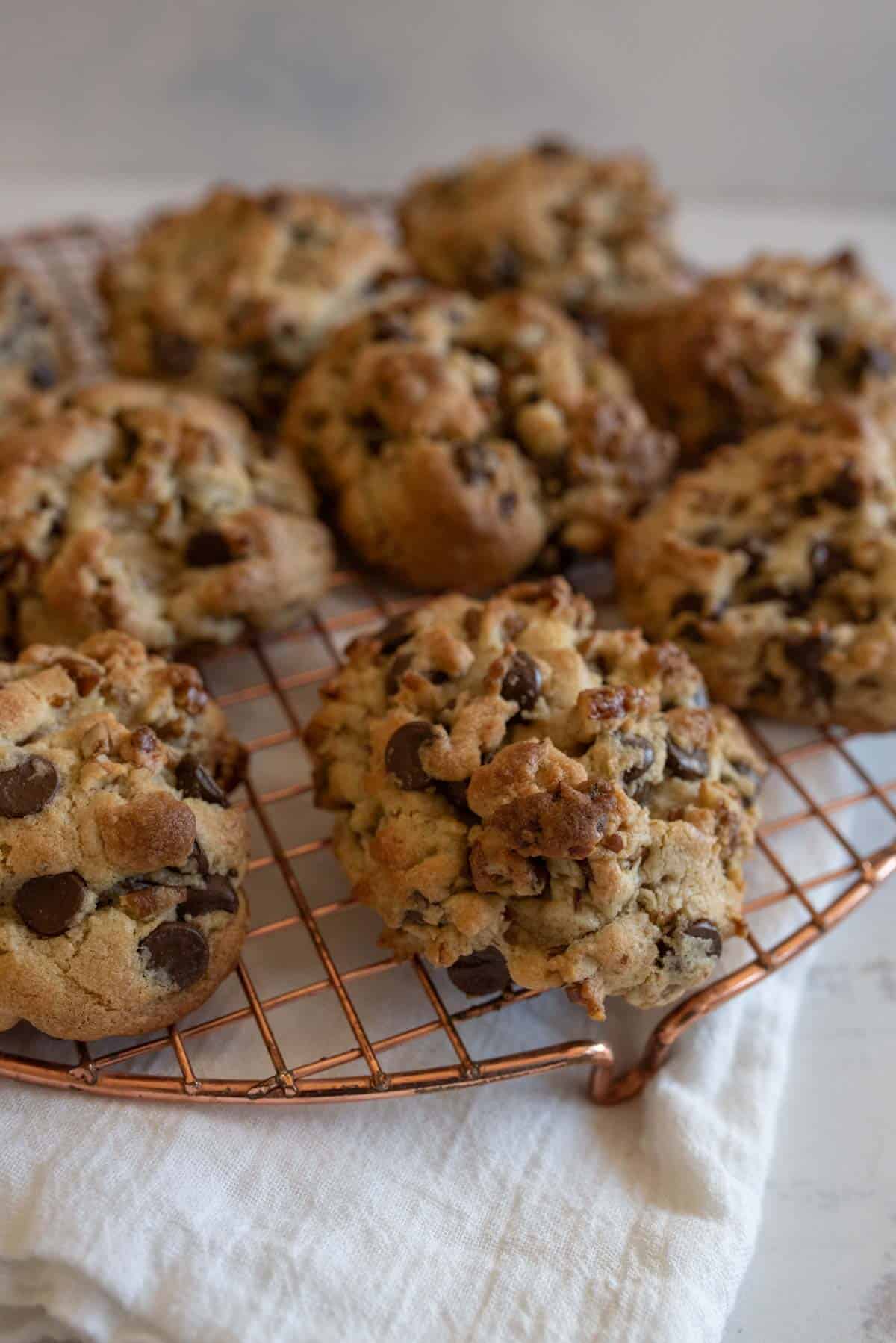 A batch of freshly baked chocolate chip cookies is cooling on a copper wire rack. The cookies are chunky, with visible chocolate chips and a golden-brown texture, placed on a white cloth on a light-colored surface.