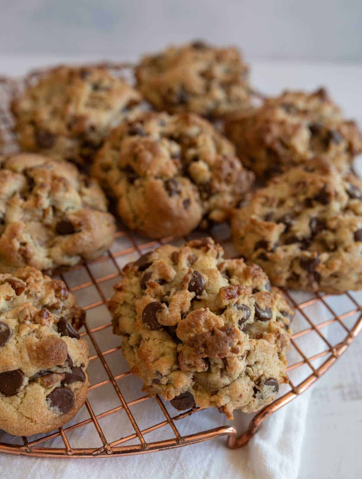 A batch of freshly baked chocolate chip cookies is cooling on a round wire rack. The cookies are chunky and packed with visible chocolate chips. They are placed on a light-colored surface.