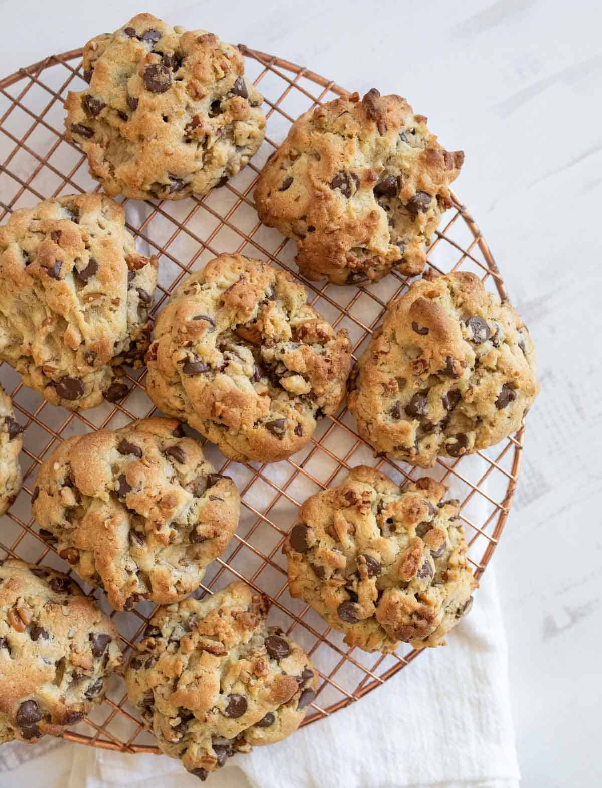 Homemade chocolate chip cookies with chunks of nuts sit on a round, gold-colored cooling rack placed on a white cloth. The cookies are unevenly shaped, with a golden-brown color, surrounded by a light, bright setting.