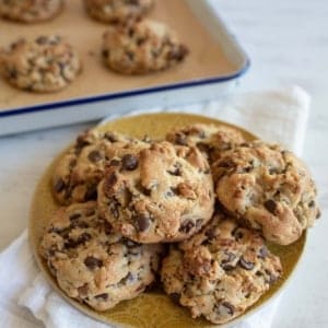 A plate piled with freshly baked chocolate chip walnut cookie recipe showing cookies sitting on a white cloth. More cookies are visible in a baking tray in the background. The cookies, based on the perfect recipe, are golden brown with visible chunks of chocolate and nuts.