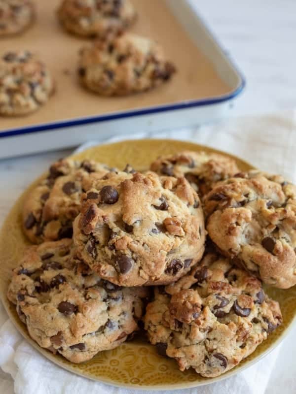 A plate piled with freshly baked chocolate chip walnut cookie recipe showing cookies sitting on a white cloth. More cookies are visible in a baking tray in the background. The cookies, based on the perfect recipe, are golden brown with visible chunks of chocolate and nuts.