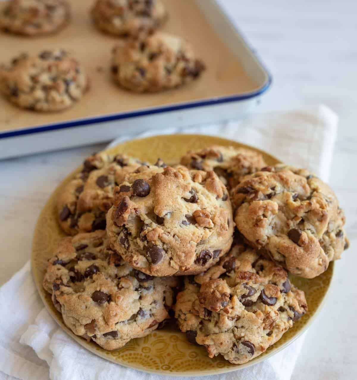 A plate piled with freshly baked chocolate chip walnut cookie recipe showing cookies sitting on a white cloth. More cookies are visible in a baking tray in the background. The cookies, based on the perfect recipe, are golden brown with visible chunks of chocolate and nuts.