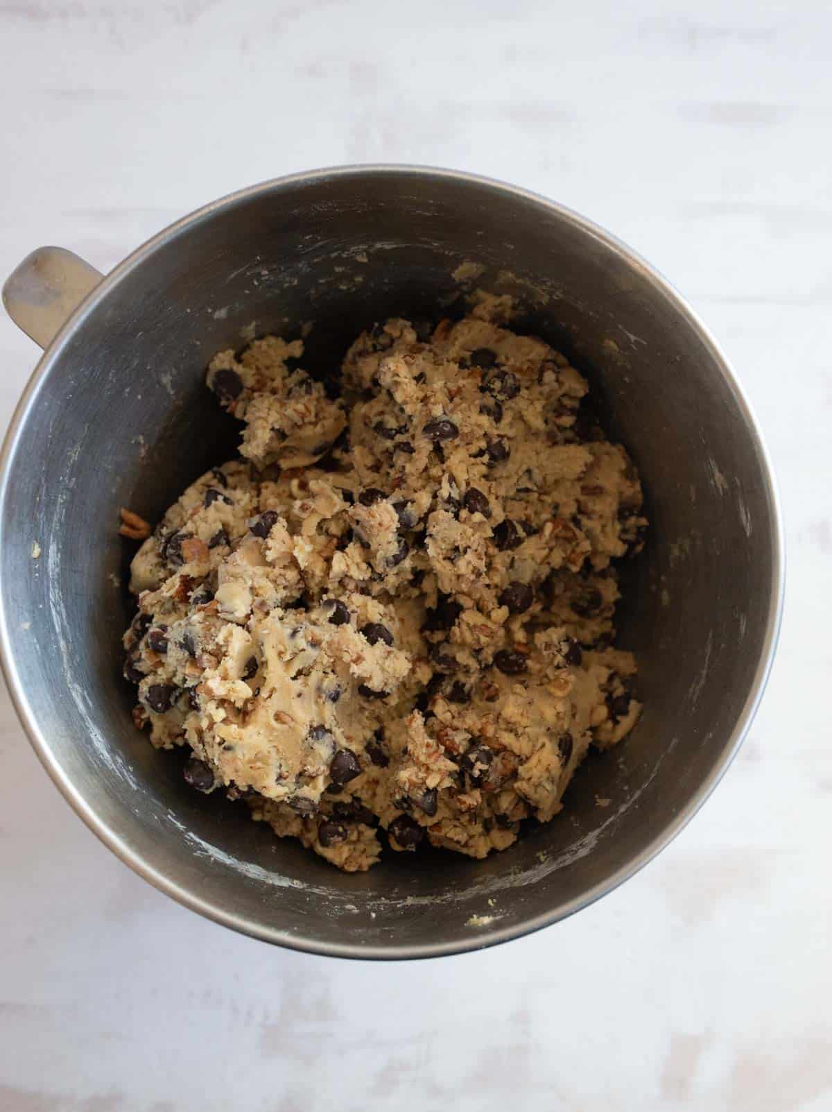 A mixing bowl filled with raw cookie dough mixed with chocolate chips and nuts, placed on a light surface.