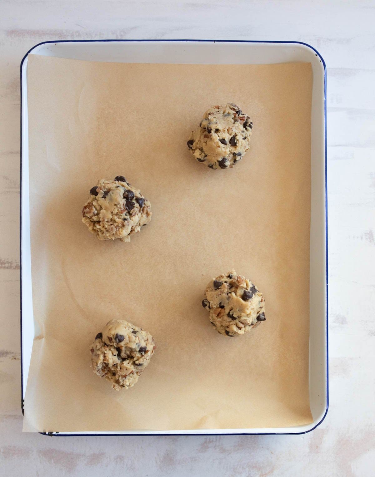 Four cookie dough balls with chocolate chips sit on parchment paper in a baking tray, ready for baking. The tray is placed on a light wooden surface.