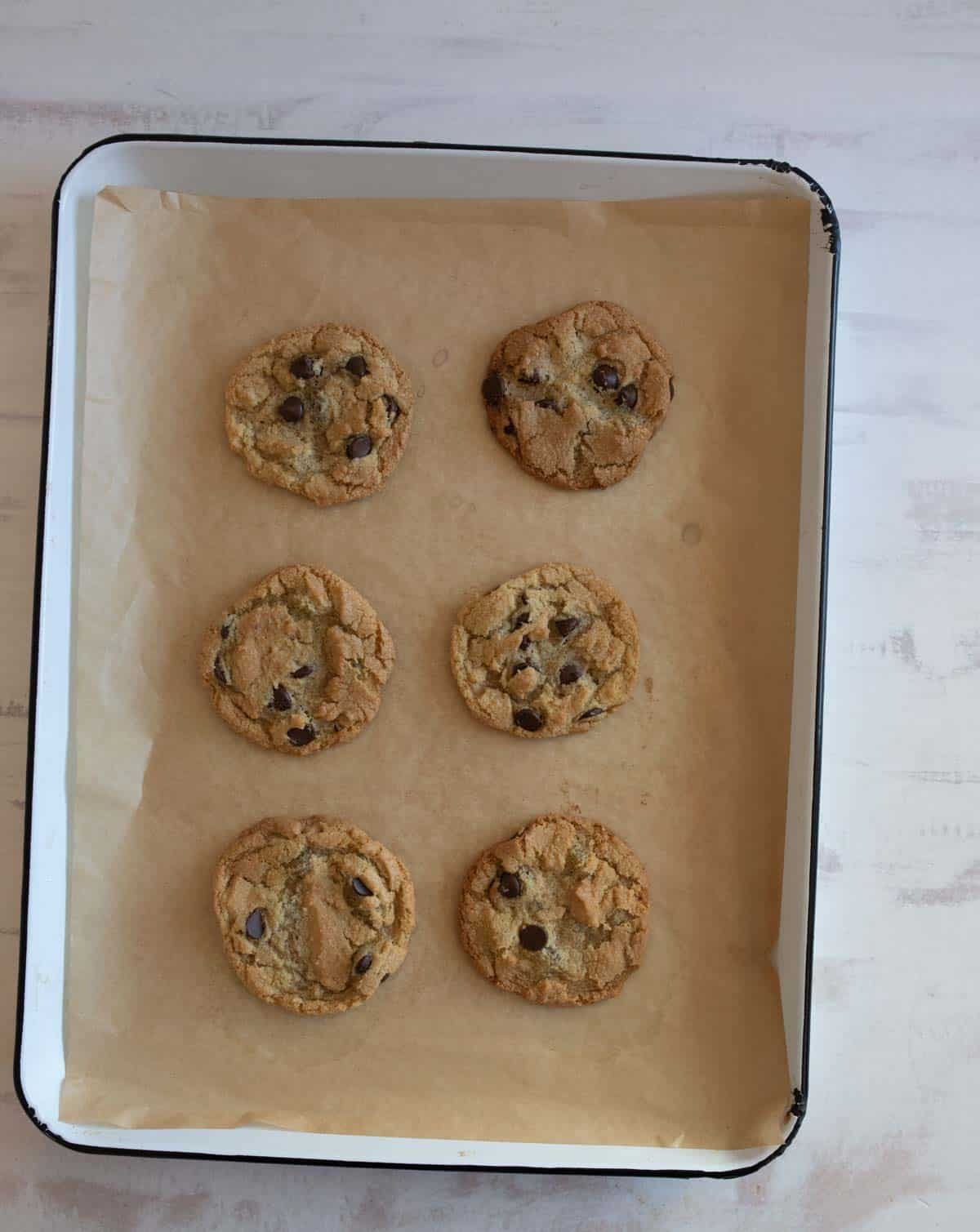 A tray of six freshly baked chocolate chip cookies on a sheet of parchment paper. The cookies are golden brown with visible chocolate chips, arranged neatly in two rows of three. The tray is set on a light-colored surface.