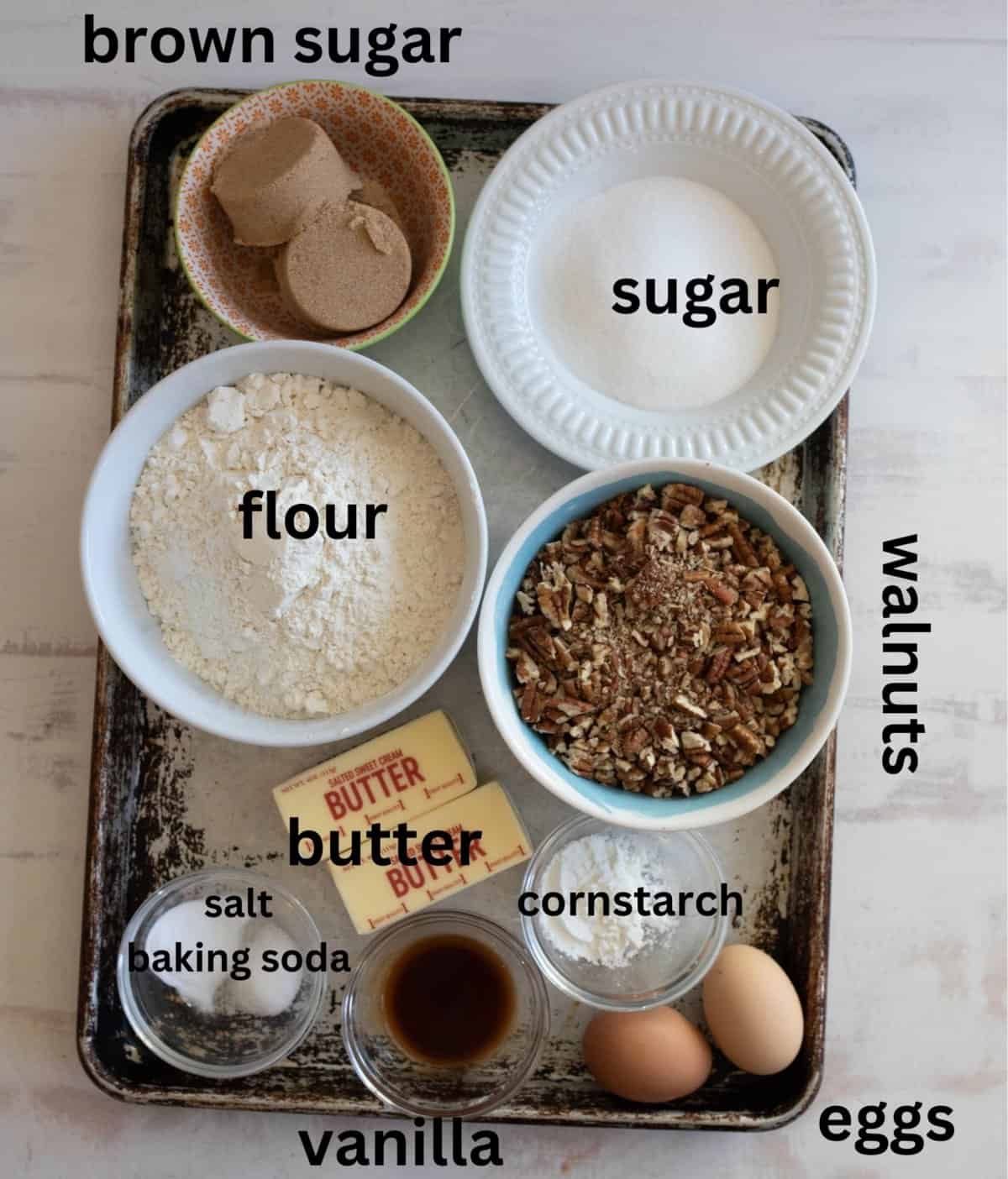 A baking tray holds labeled ingredients: brown sugar, sugar, flour, walnuts, butter, eggs, vanilla, salt, baking soda, and cornstarch, all in separate containers.