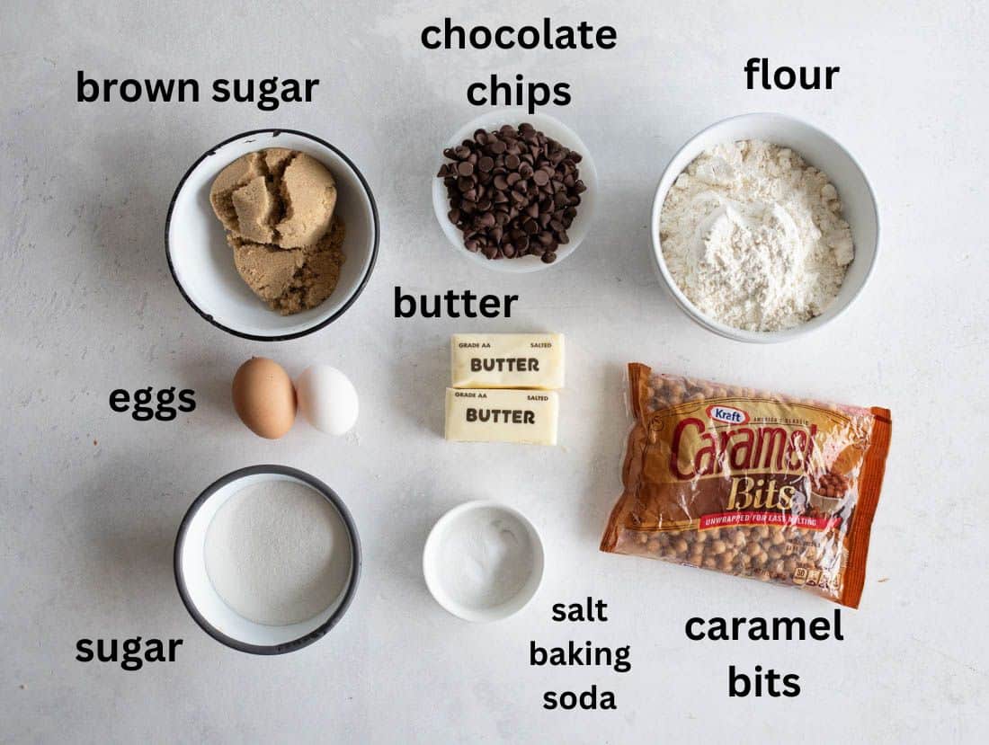 Ingredients for baking displayed on a light surface: bowls of brown sugar, chocolate chips, flour, sugar, salt, and baking soda; two eggs; sticks of butter; a bag of caramel bits. Labels identify each item.