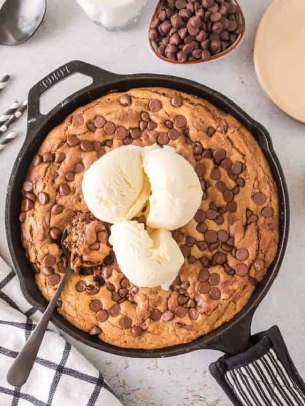 Cast iron chocolate chip cookies topped with chocolate chips and three scoops of vanilla ice cream. A small chunk is scooped out with a spoon, revealing gooey goodness. Surrounding items include a dish of chocolate chips, a striped towel, and a partial view of plates.