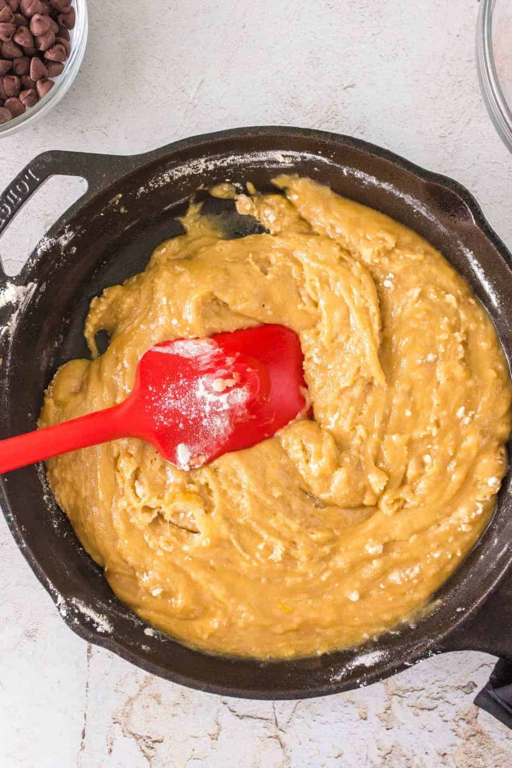 Batter for cast iron chocolate chip cookies is being mixed in a black bowl with a red spatula. The mixture appears smooth and creamy, resting on a light-colored countertop. A small bowl of chocolate chips is partially visible in the top left corner, ready to be added to the delicious creation.