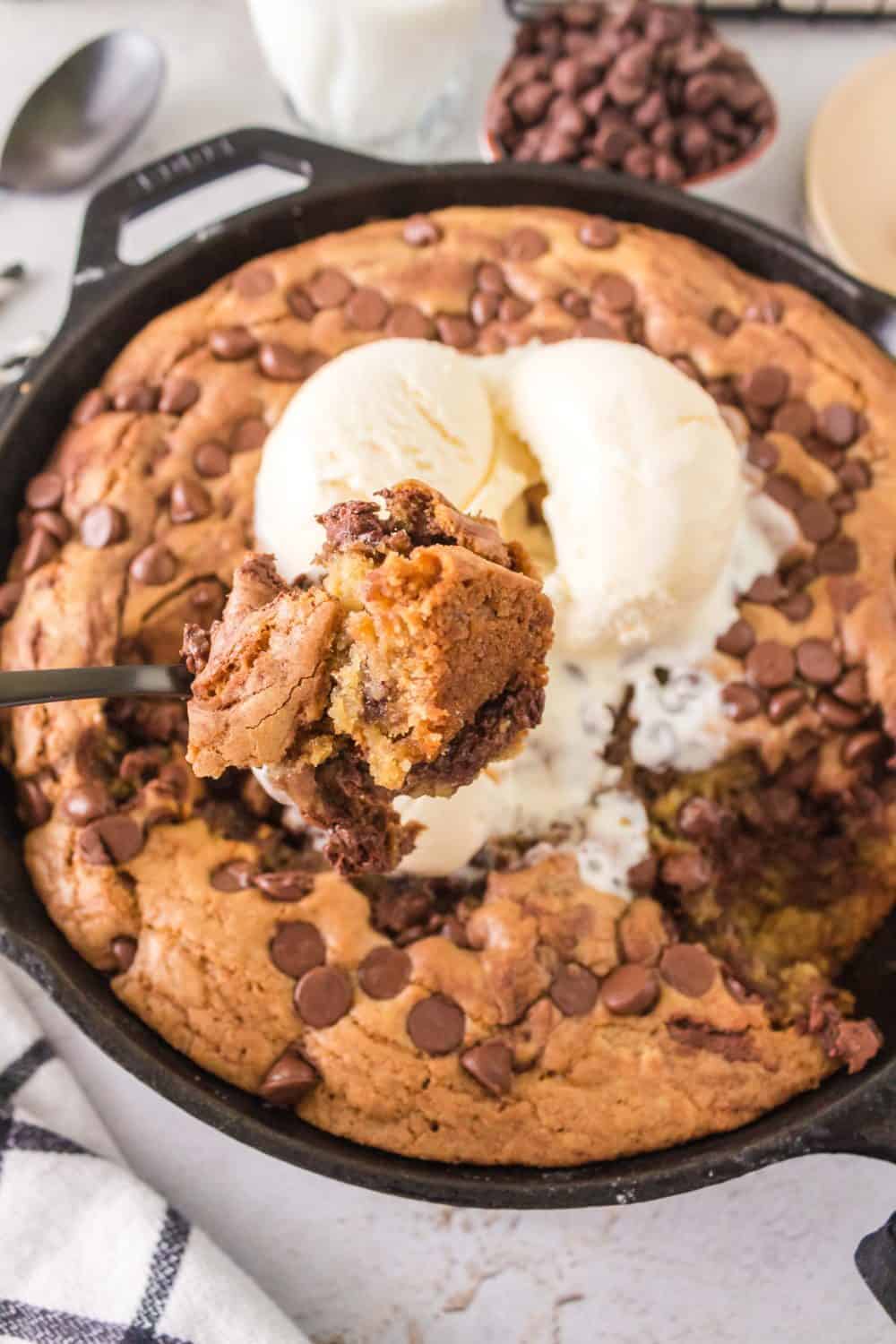 A skillet cookie, baked in a cast iron pan, is topped with vanilla ice cream and chocolate chips. A spoon holds a portion of the gooey treat. Chocolate chips are scattered in the background, with a checkered napkin partially visible, adding to the cozy charm.