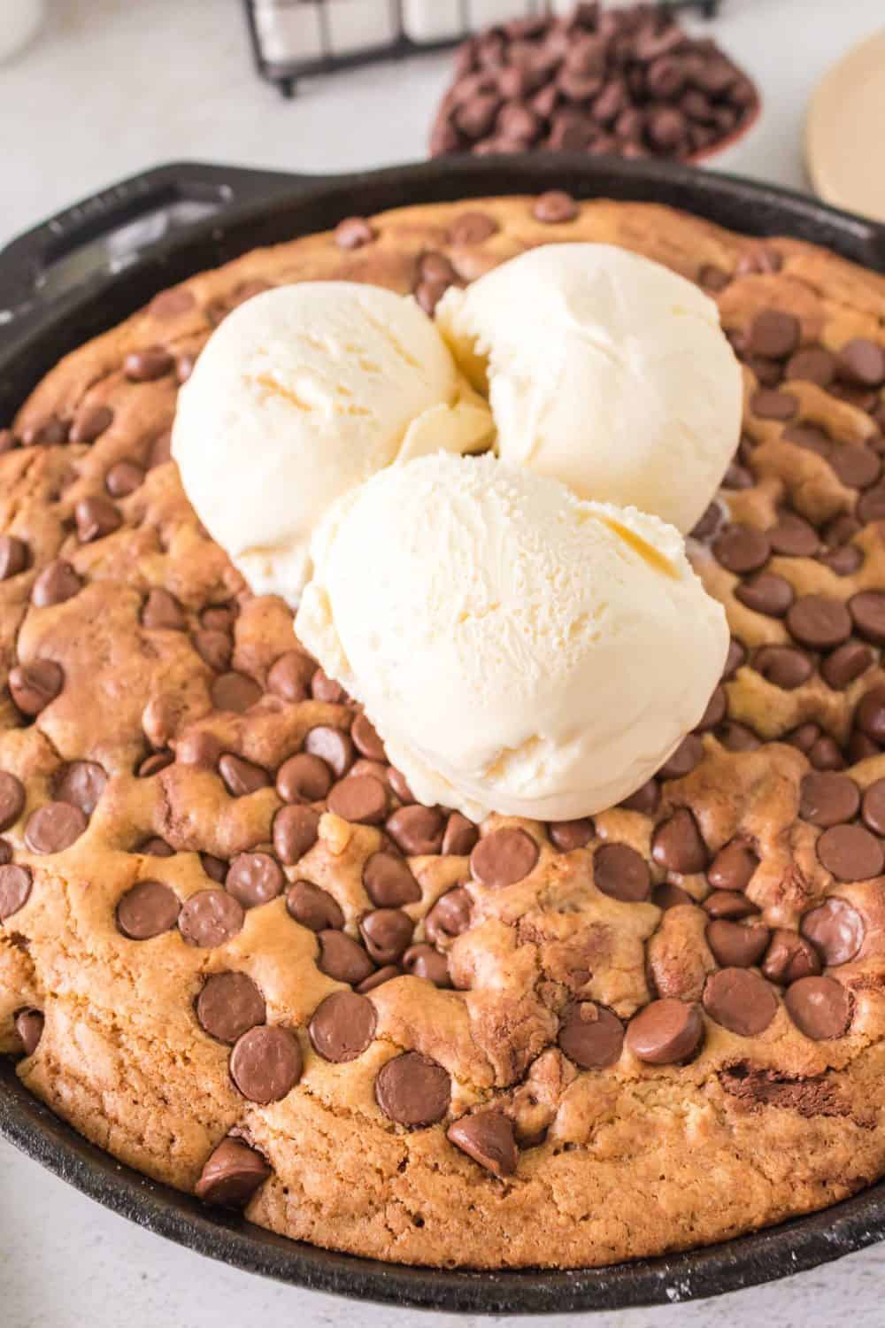 A large cast iron chocolate chip cookie is baked to perfection and topped with three sumptuous scoops of vanilla ice cream. Additional chocolate chips are scattered over the cookie surface, with a bowl of extra chips visible in the background for a delightful touch.