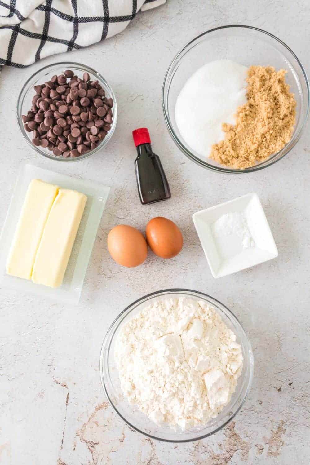 Baking ingredients arranged on a light surface: chocolate chips for cast iron chocolate chip cookies in a bowl, white and brown sugar in another, a small bottle of vanilla extract, two sticks of butter, two eggs, a bowl of flour, and a small dish with baking soda. A checkered cloth nearby.