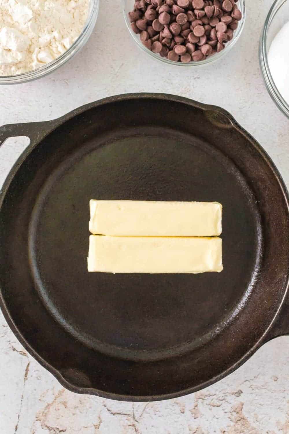 Two sticks of butter rest side by side in a black cast-iron skillet, ready to create delicious cast iron chocolate chip cookies. Around the skillet, glass bowls hold chocolate chips, flour, and sugar on a light countertop, setting the stage for a sweet baking adventure.