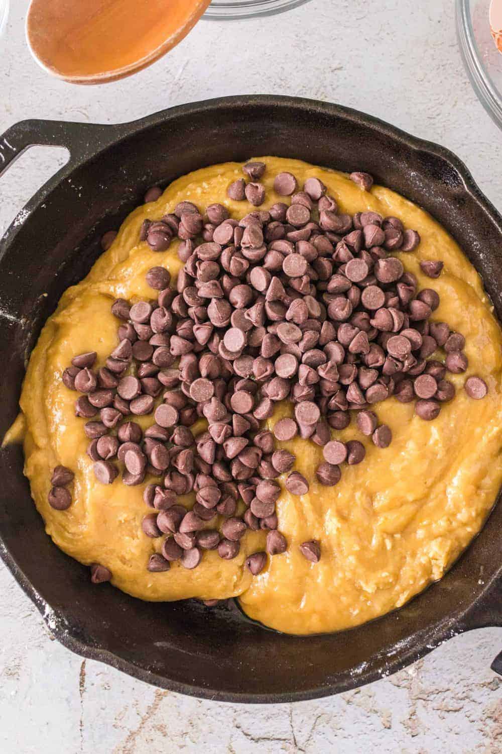 A cast iron skillet brims with raw cookie dough topped with chocolate chips, resting on a light-colored countertop. A wooden spoon peeks into the scene from the top left corner.