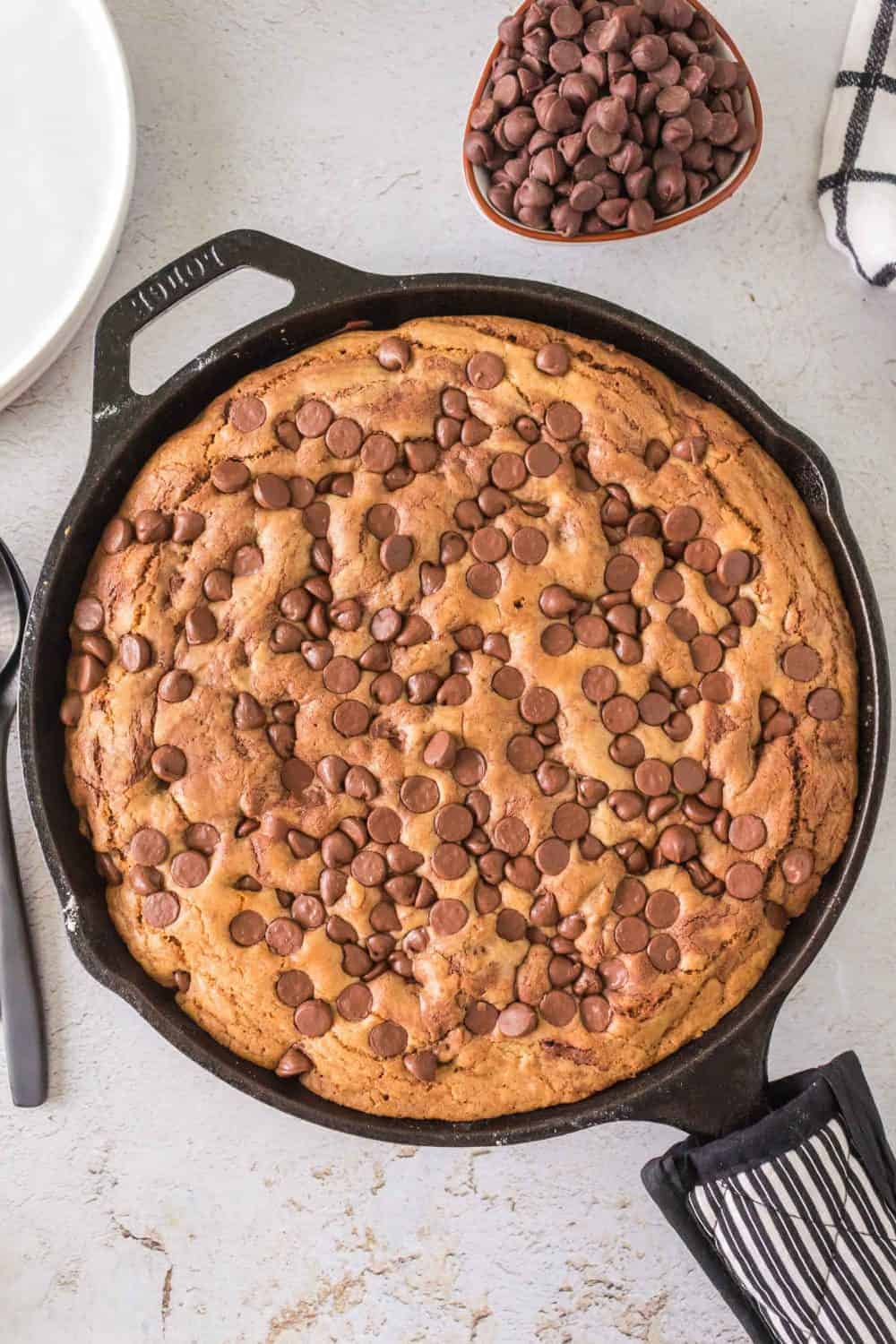 A large skillet cookie brimming with chocolate chips rests on a light surface. Crafted as cast iron chocolate chip cookies, its accompanied by a small bowl overflowing with more chocolate chips, a white plate, and a black spoon. A black and white striped cloth is partially visible in the scene.