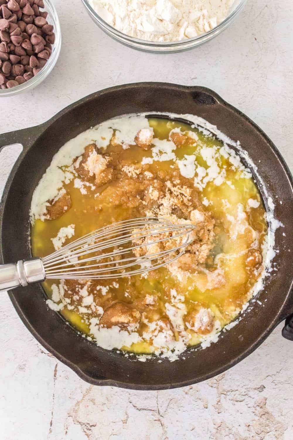 A top view of a cast iron skillet containing a mixture of cream, sugar, and melted butter being whisked together. Surrounding the skillet are bowls filled with chocolate chips and flour on a light-textured countertop, setting the stage for delectable chocolate chip cookies.