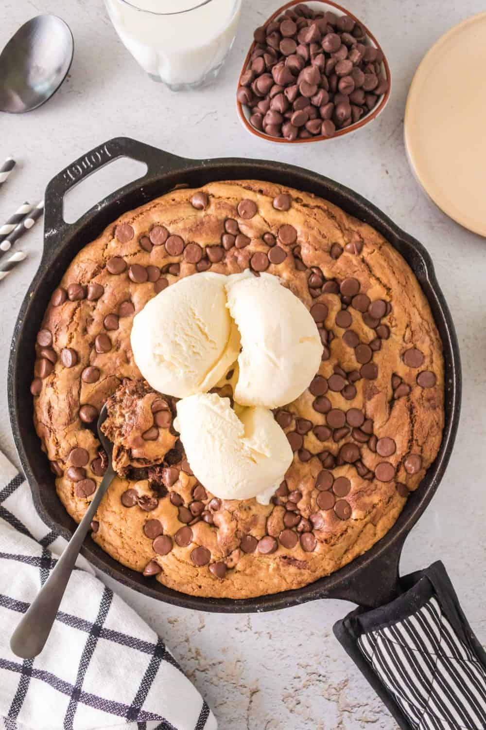 A cast iron chocolate chip cookie is topped with three scoops of vanilla ice cream. A spoon rests in the cookie, surrounded by a black oven mitt, checked towel, plate, spoon, and bowl of chocolate chips on a light surface. A glass of milk stands invitingly in the background.