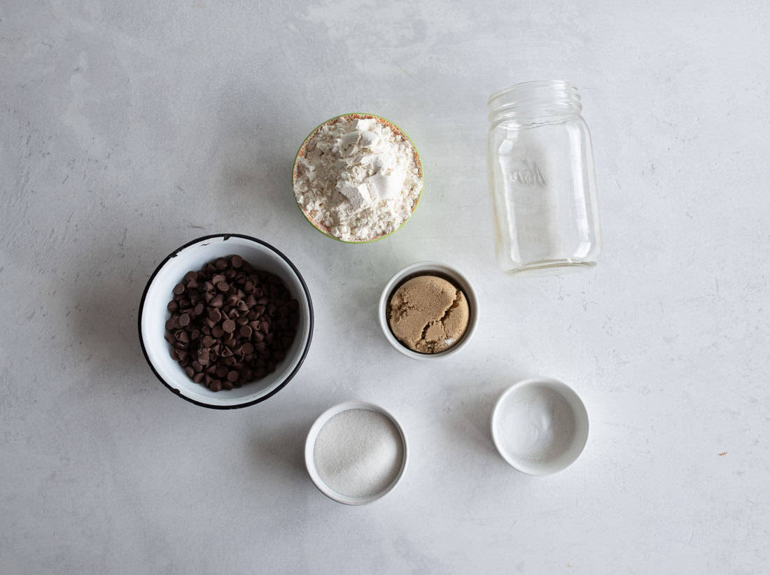 Ingredients for baking include a bowl of chocolate chips, a bowl of flour, an empty glass jar, a bowl of brown sugar, a bowl of white sugar, and a bowl of salt, all arranged on a light gray surface.