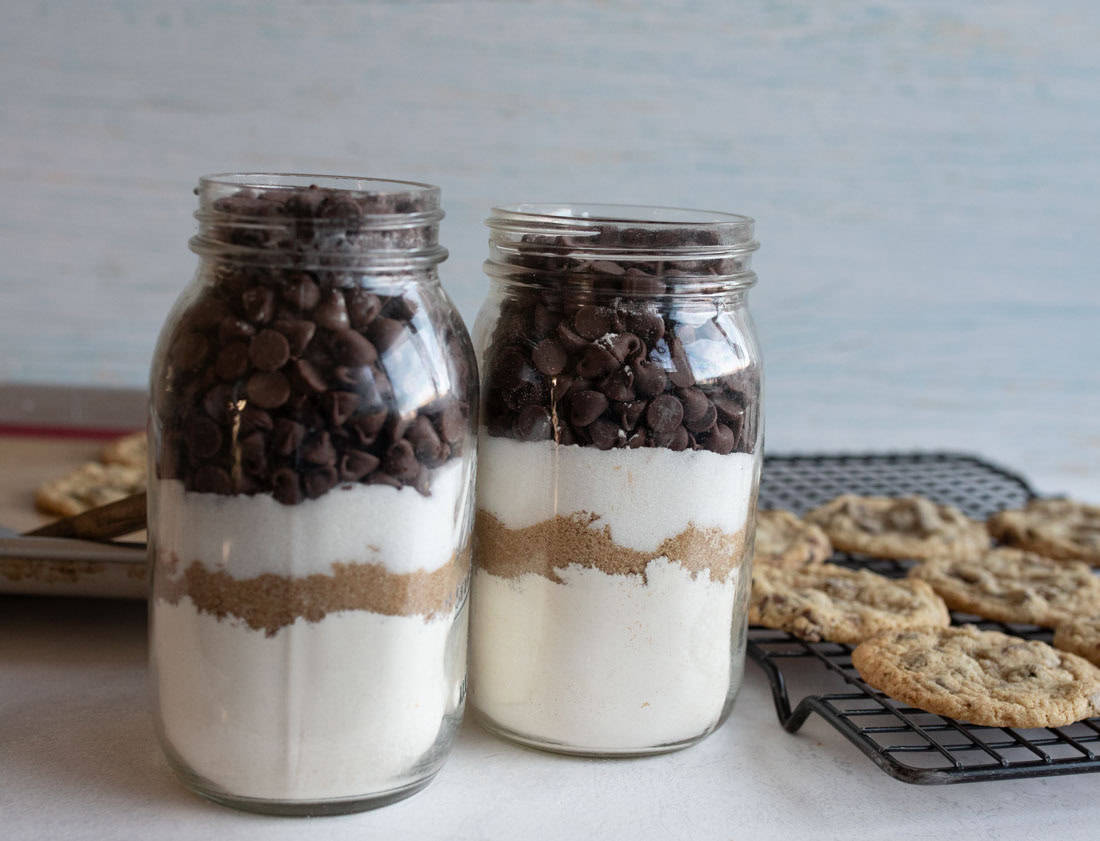 Two glass jars filled with layered ingredients for cookies, including flour, brown sugar, and chocolate chips, are placed on a table. Baked cookies are cooling on a wire rack in the background.