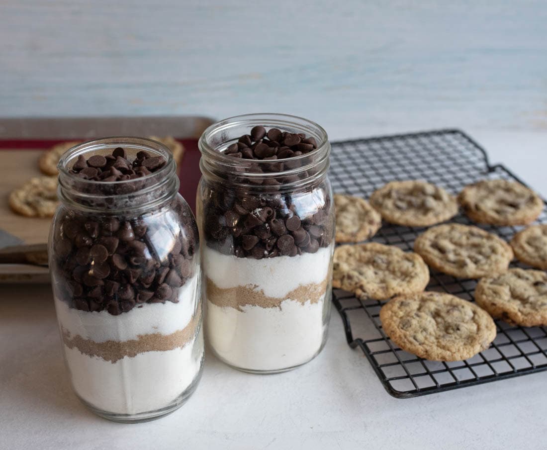 Two glass jars filled with layered cookie mix ingredients, topped with chocolate chips, are on a countertop. A cooling rack with freshly baked cookies is in the background.