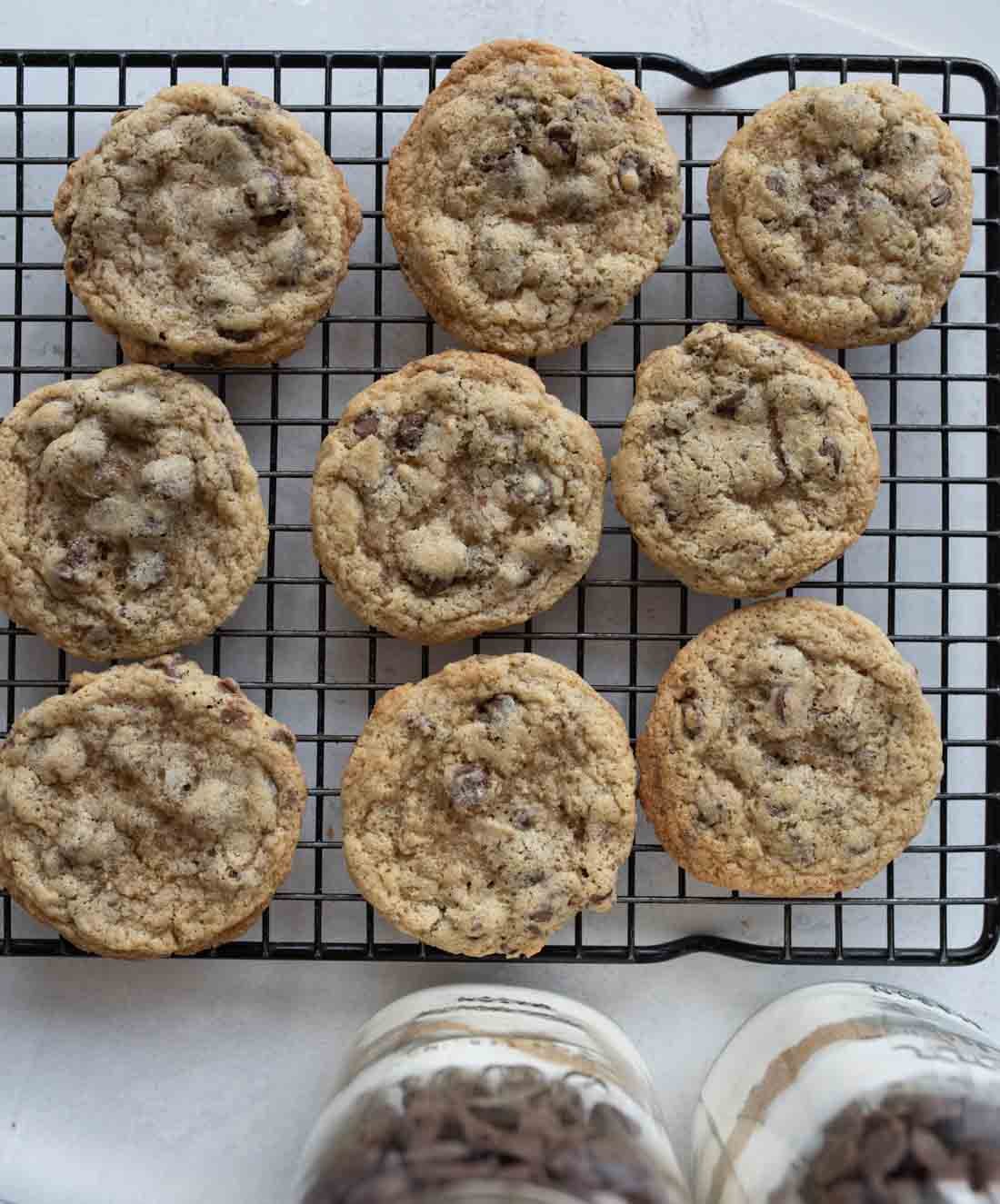 Nine chocolate chip cookies cooling on a black wire rack. Below the rack, there are jars filled with chocolate chips and flour. The cookies are golden brown with visible chocolate chips.