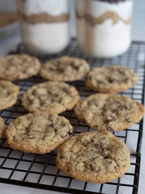 Chocolate chip cookies cooling on a wire rack, with two jars of layered cookie ingredients in the background. The cookies are golden brown and freshly baked from a mason jar chocolate chip cookie recipe.