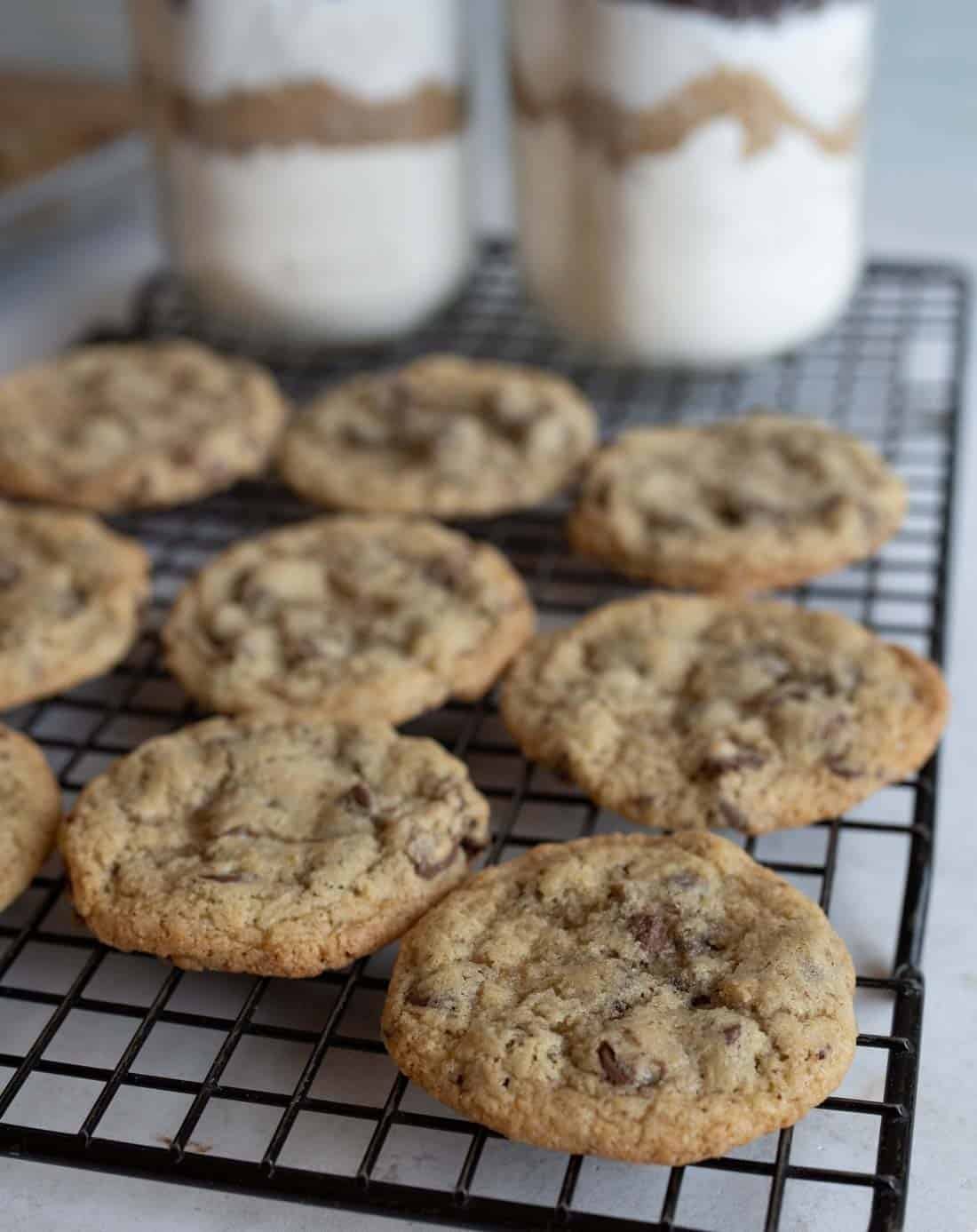 Chocolate chip cookies cooling on a wire rack, with two jars of layered cookie ingredients in the background. The cookies are golden brown and freshly baked from a mason jar chocolate chip cookie recipe.