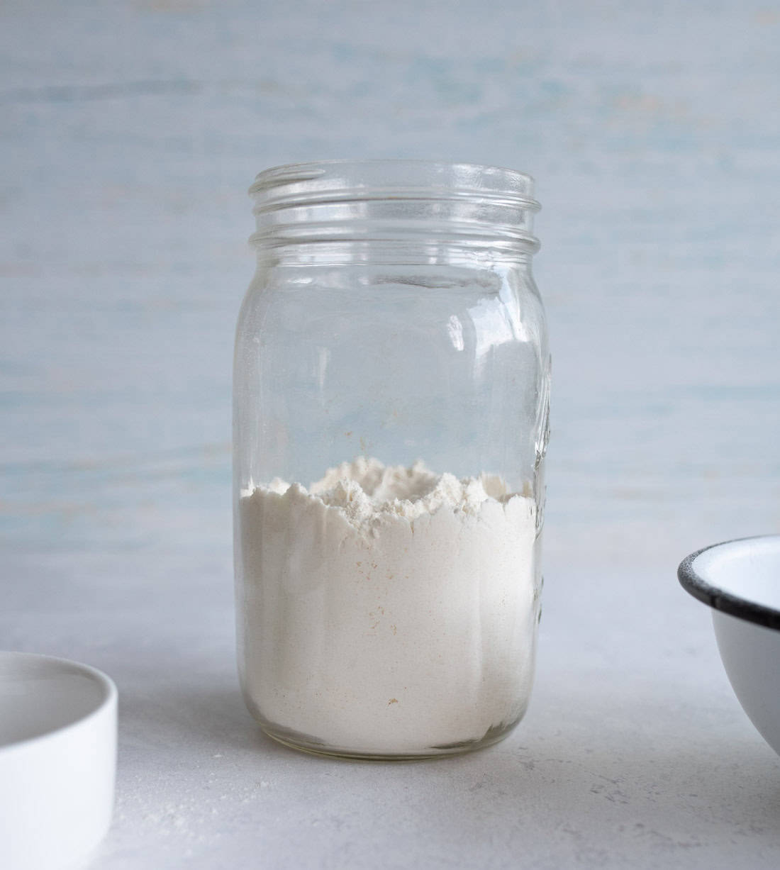 A glass jar filled with white flour sits on a light gray surface. A portion of a white ceramic dish is visible on the bottom left, and a metal-rimmed bowl appears partially on the right. The background is a faded blue.