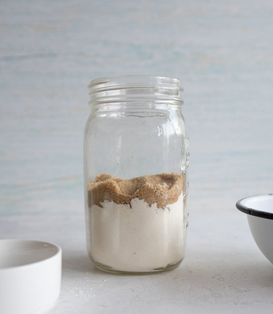 A glass jar is filled with layered white and brown powders, likely sugar. It is placed on a light-colored tabletop with blurred bowls nearby. The background is softly textured with pale blue and white hues.