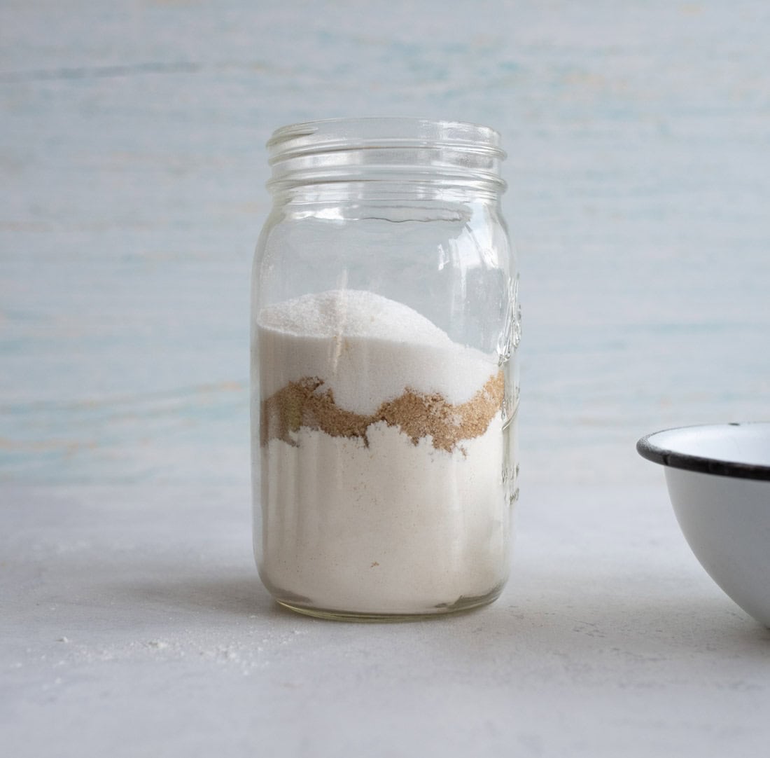 A clear glass jar filled with layers of different sugars sits on a light surface. The background is a pale blue-gray texture. A partial view of a white bowl with a dark rim is visible on the right.