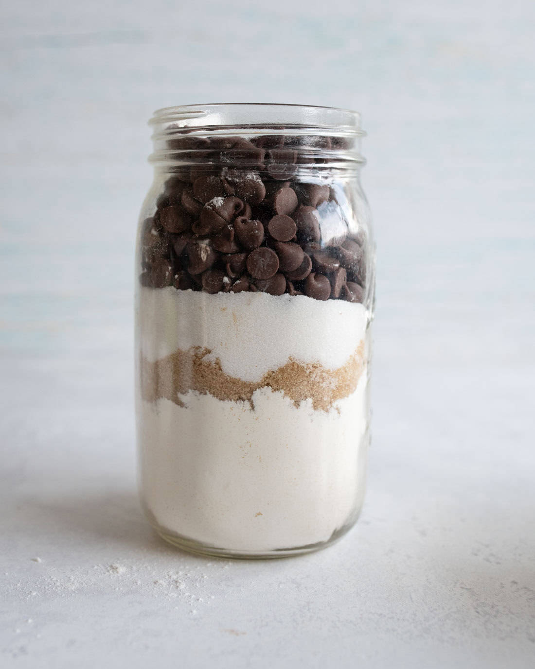 A glass jar filled with layered baking ingredients: chocolate chips on top, followed by brown sugar, and white flour at the bottom, against a light background.