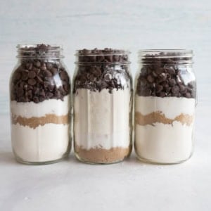 Three glass jars filled with layers of baking ingredients: chocolate chips, flour, white sugar, and brown sugar, standing on a light surface with a neutral background.